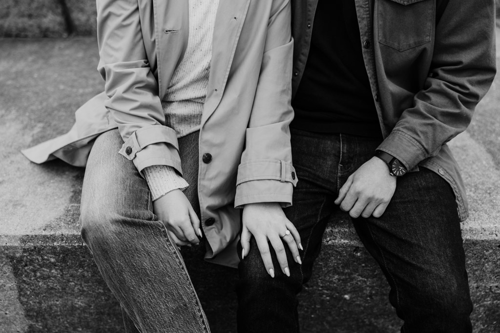 Black and white photo of engaged couple sitting close together on concrete steps at Belmont Harbor shown from the shoulders down with their hands and engagement ring visible