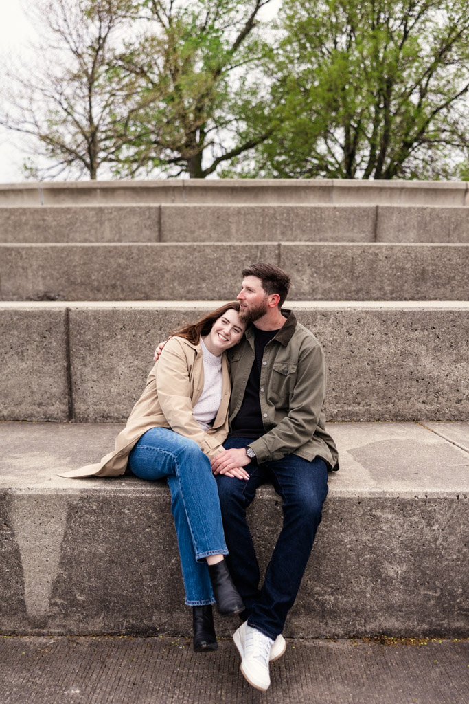 A couple sits on concrete steps at Belmont Harbor, smiling and cuddling during their sunrise engagement session, with trees in the background