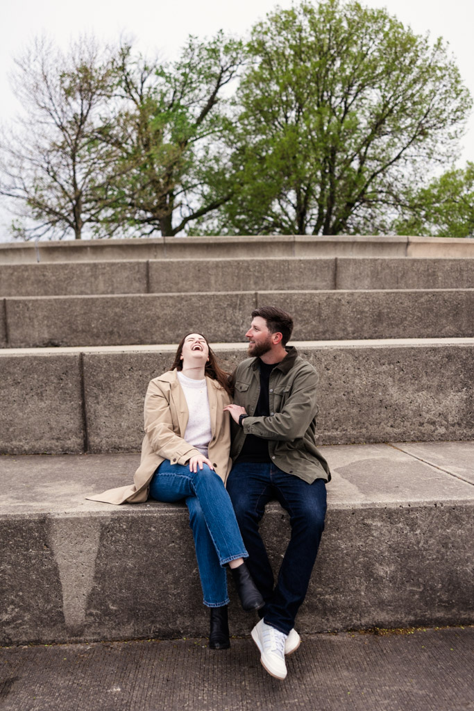 happy couple sits on concrete steps at Belmont Harbor, sharing a laugh together in the early light of their sunrise engagement session with trees in the background