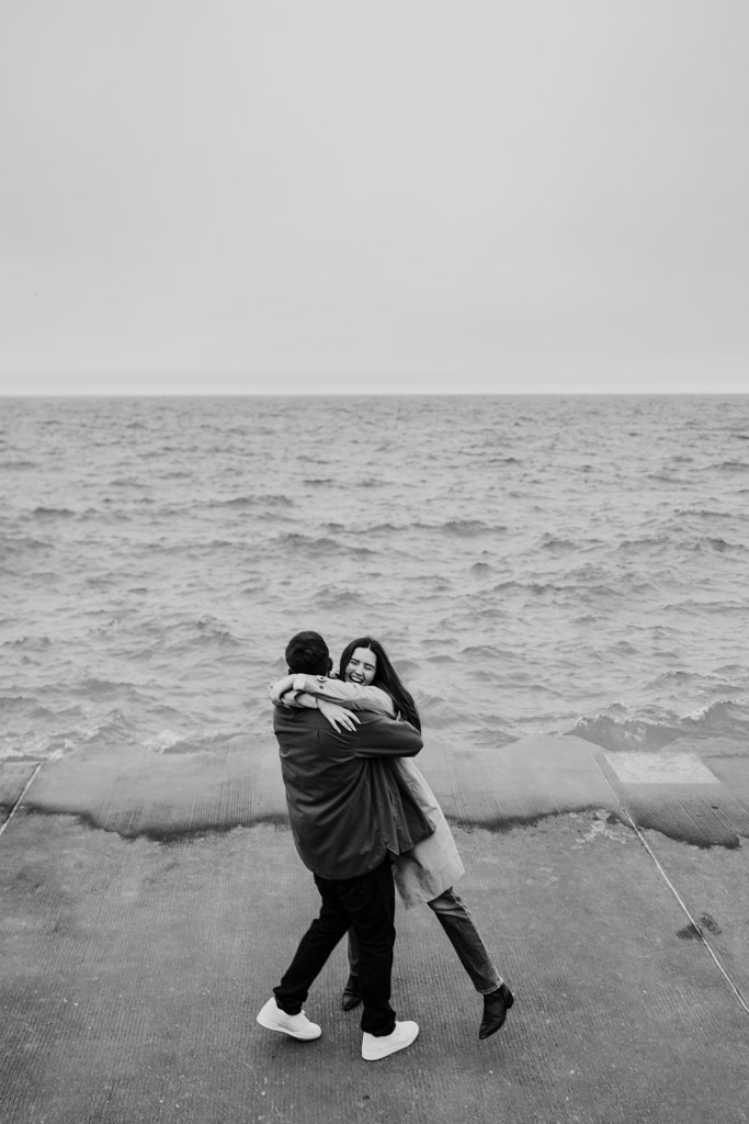 A couple embraces and laughs by Lake Michigan during their sunrise Belmont Harbor engagement session, with waves and a cloudy sky in the background