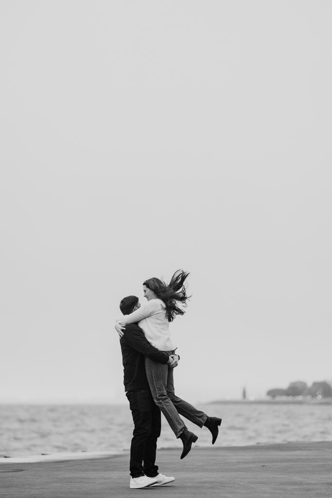 At sunrise, a man lifts his fiancée by Lake Michigan on a cloudy day during their Belmont Harbor engagement session