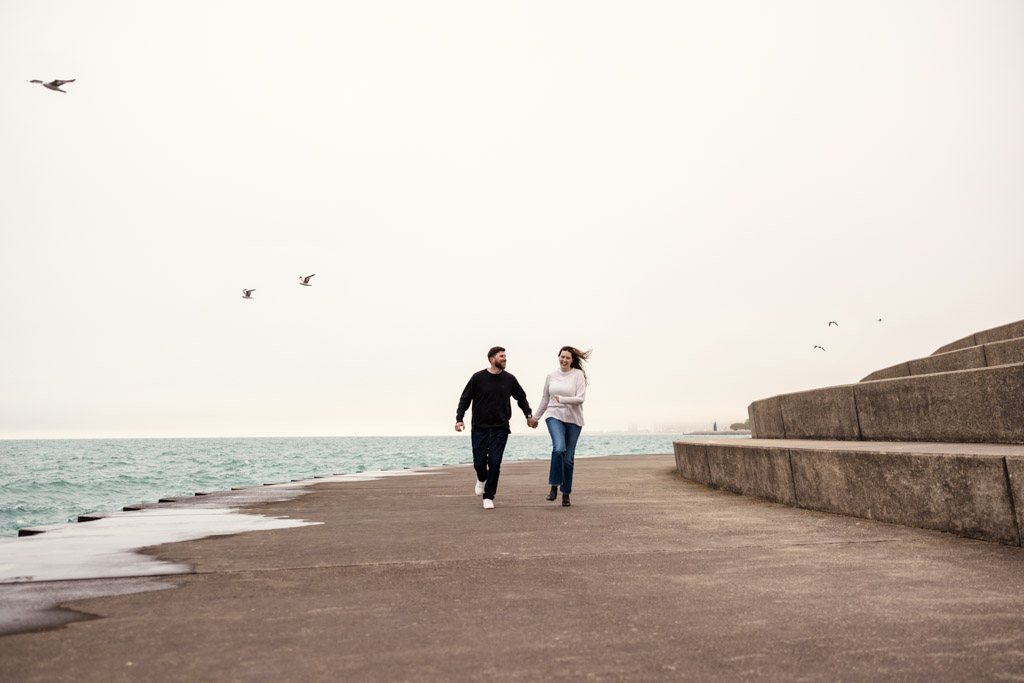 Engaged couple holding hands walks by Lake Michigan at sunrise on a cloudy day, with seagulls flying overhead