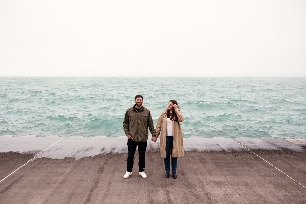 A couple stands holding hands on a concrete pier at Belmont Harbor by Lake Michigan at sunrise