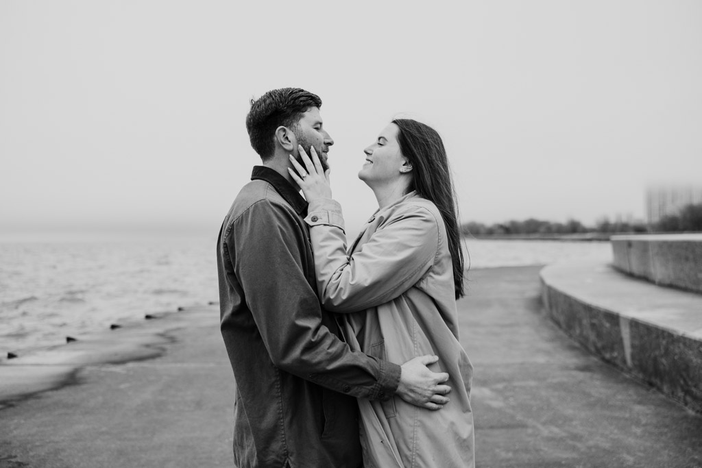 Black and white photo of couple holding each other standing by Lake Michigan at Belmont Harbor at sunrise