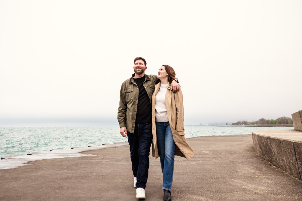 Happy engaged couple walks by Lake Michigan at Belmont Harbor on a cloudy day, smiling and gazing at each other