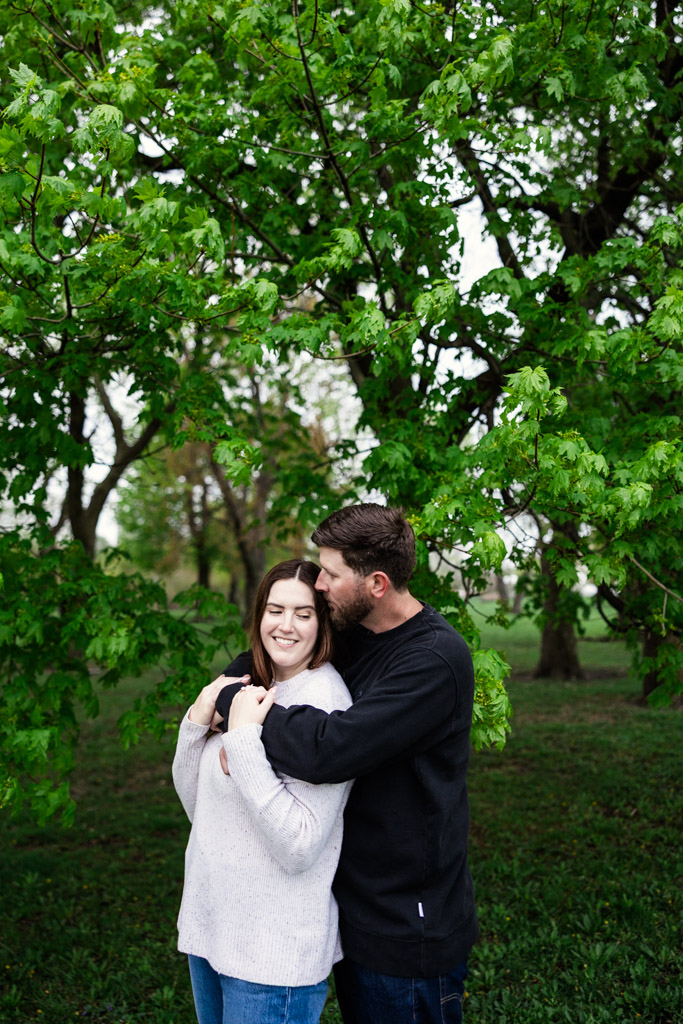 A couple stands outdoors at Belmont Harbor during their sunrise engagement session; the man hugs the smiling woman from behind under green leafy trees.