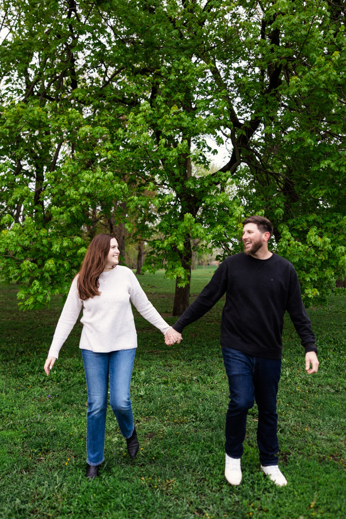 Engaged couple holding hands and walking on grass in Belmont Harbor with green trees