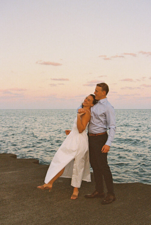 A couple laughs together by the ocean at sunset, standing on a pier with a pastel sky behind them—capturing a Best of 2025 moment.