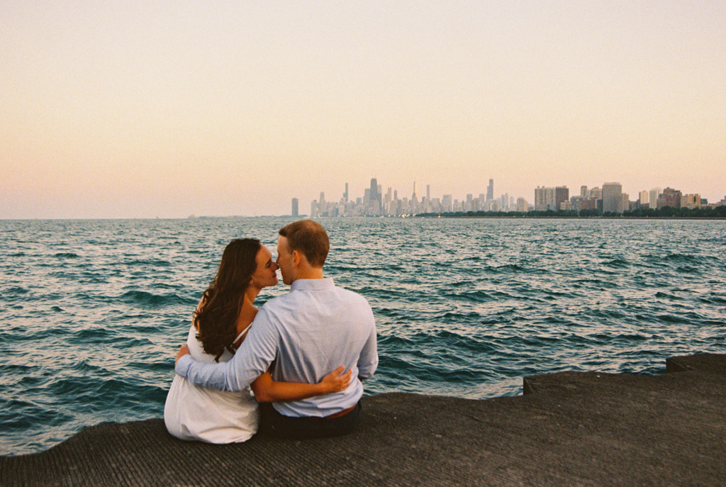 Film photo of engaged couple sitting by Lake Michigan with Chicago skyline in the distance at sunset