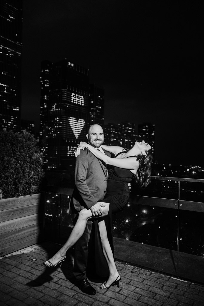 Black and white photo of smiling couple poses on a rooftop at night with city lights and a heart shape on a building behind them during their Cindy's Rooftop wedding reception
