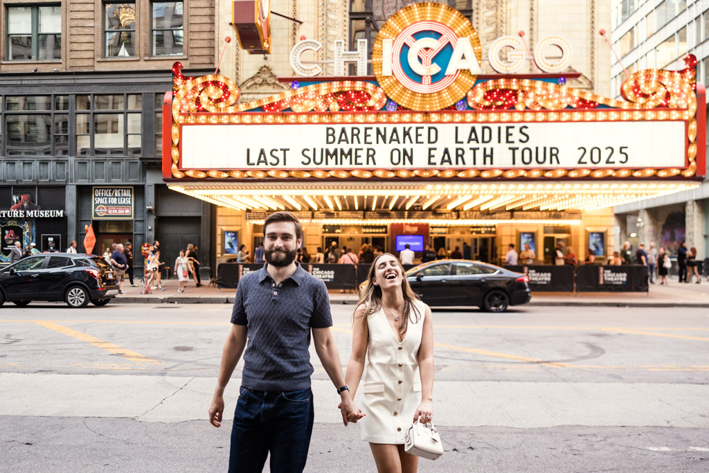 A smiling couple holds hands in front of the Chicago Theatre marquee during their summer engagement session