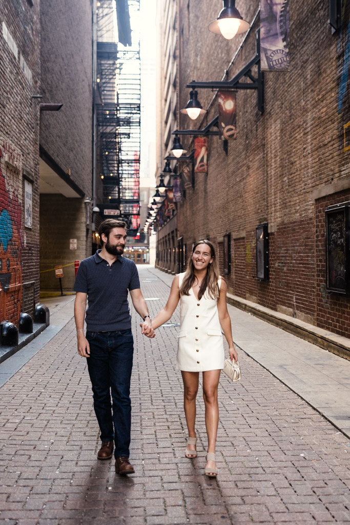 A couple holding hands and smiling while walking down a brick alleyway with murals and hanging lights during their Chicago summer engagement session