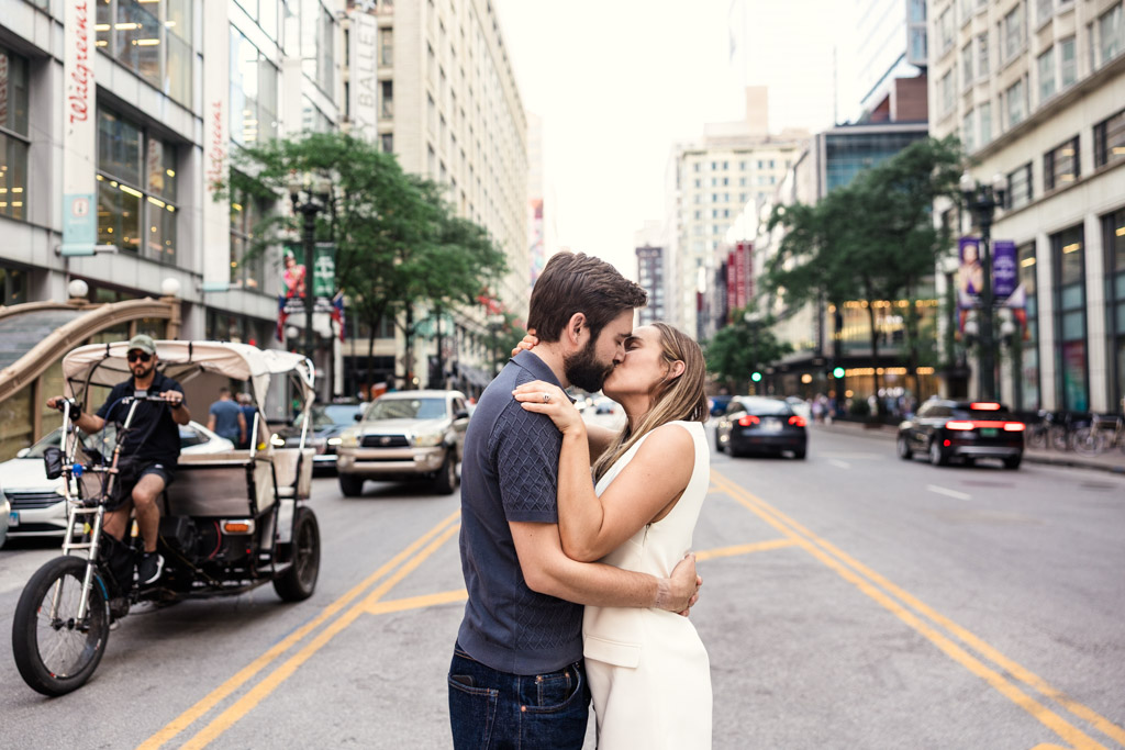 Couple kisses in the middle of a downtown Chicago street, surrounded by traffic and buildings during their summer engagement session