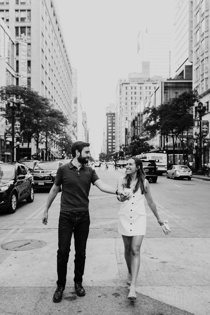 Black and white photo of happy engaged couple holding hands while walking down a busy Chicago street, surrounded by cars and tall buildings
