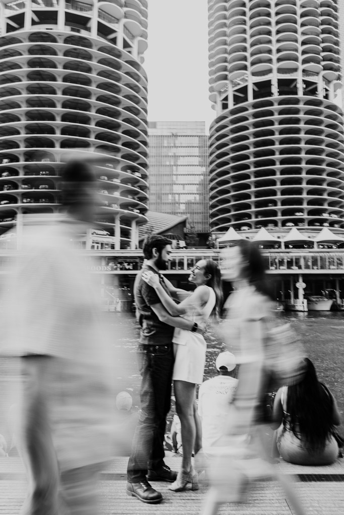 Creative black and white photo of couple holding each other on the Chicago Riverwalk during their summer engagement session, with blurred people walking and tall buildings in the background