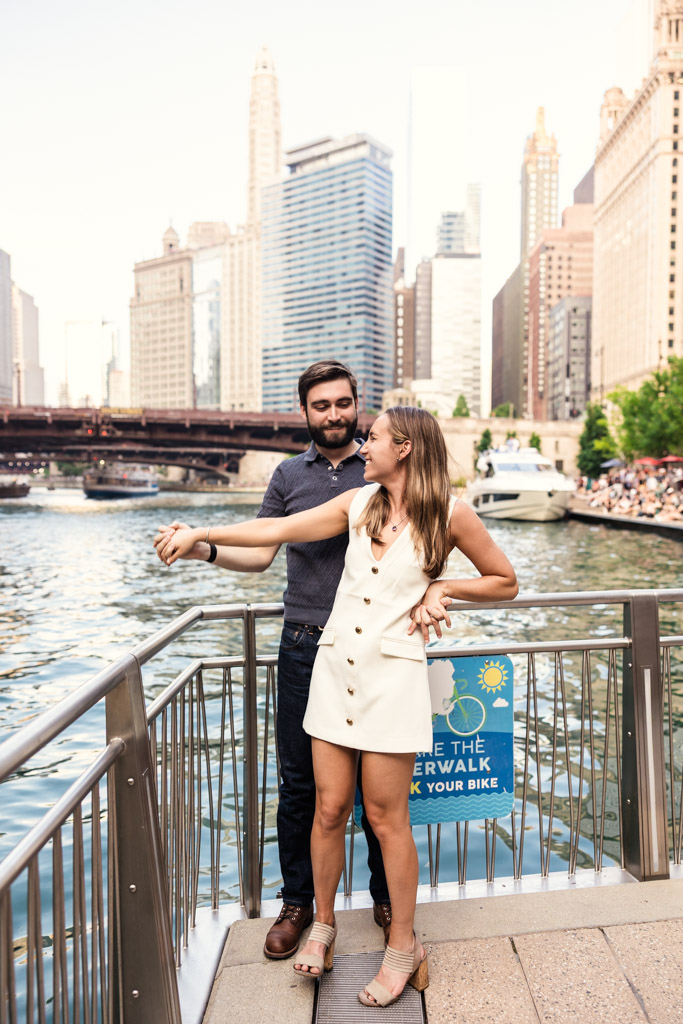 Happy couple stands on the Chicago Riverwalk during their Chicago summer engagement sessions, with city buildings and a bridge in the background