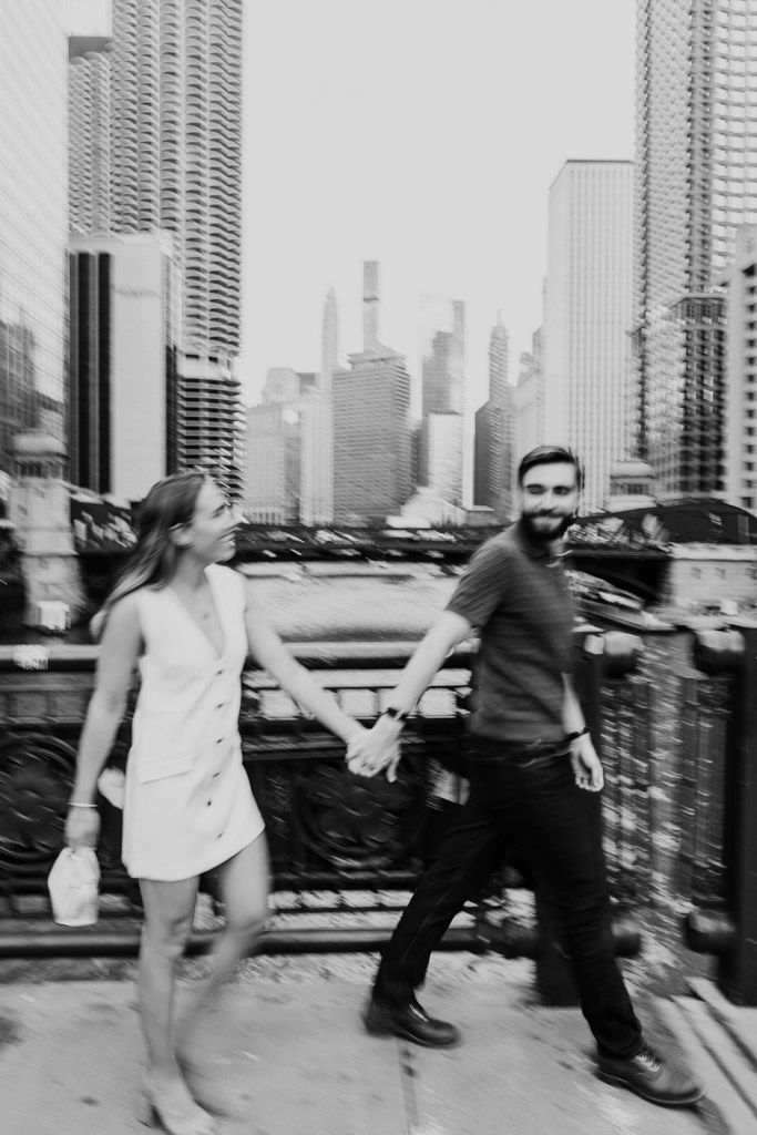 Creative motion-blur photo of couple holding hands and walking on LaSalle Street Bridge in Chicago, with tall buildings in the background during their summer engagement session
