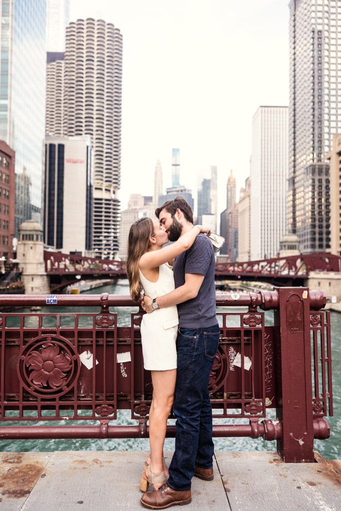 A couple kisses on LaSalle Street Bridge in Chicago with tall buildings in the background during their Chicago summer engagement session