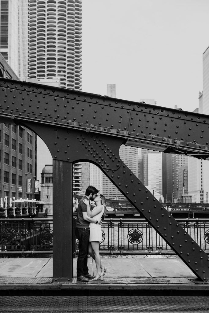 Black and white photo of engaged couple kissing on LaSalle Street Bridge with downtown Chicago skyscrapers in the background