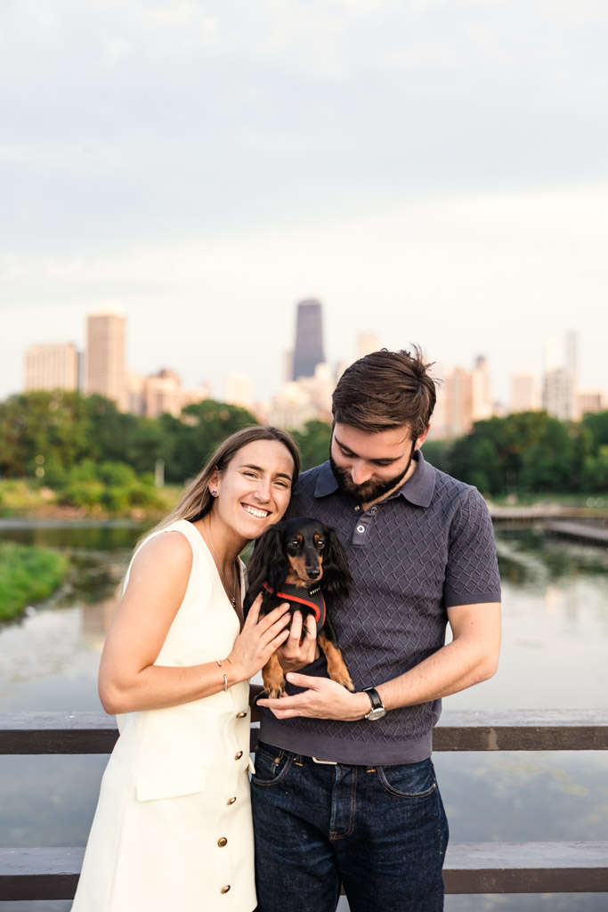 Happy couple poses with their dachshund on a Lincoln Park bridge during their Chicago summer engagement session, the city skyline visible in the background