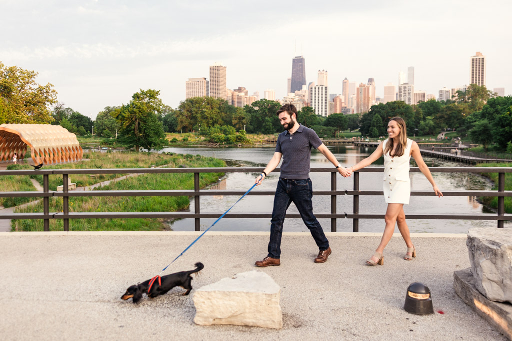A couple holding hands walks a small black dachshund on a leash in Lincoln Park with the Chicago skyline in the background during their summer engagement session