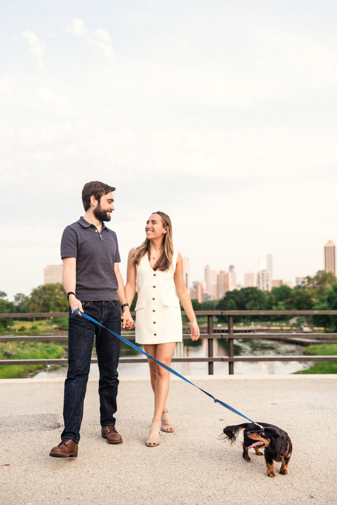 Engaged couple holding hands walks a small dog on a leash in Lincoln Park with the Chicago skyline in the background