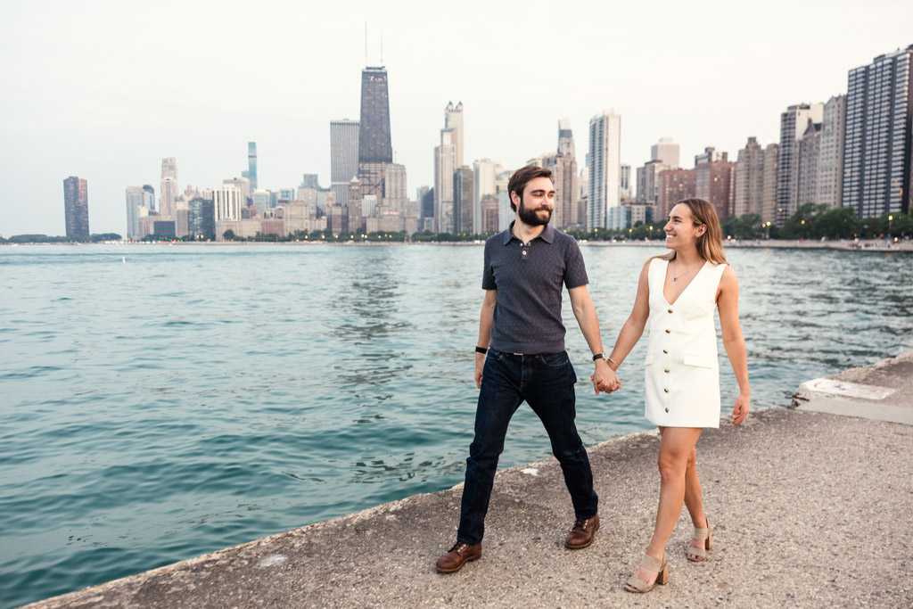 A couple walks hand in hand along the lakefront during a Chicago summer engagement session, with the city skyline in the background