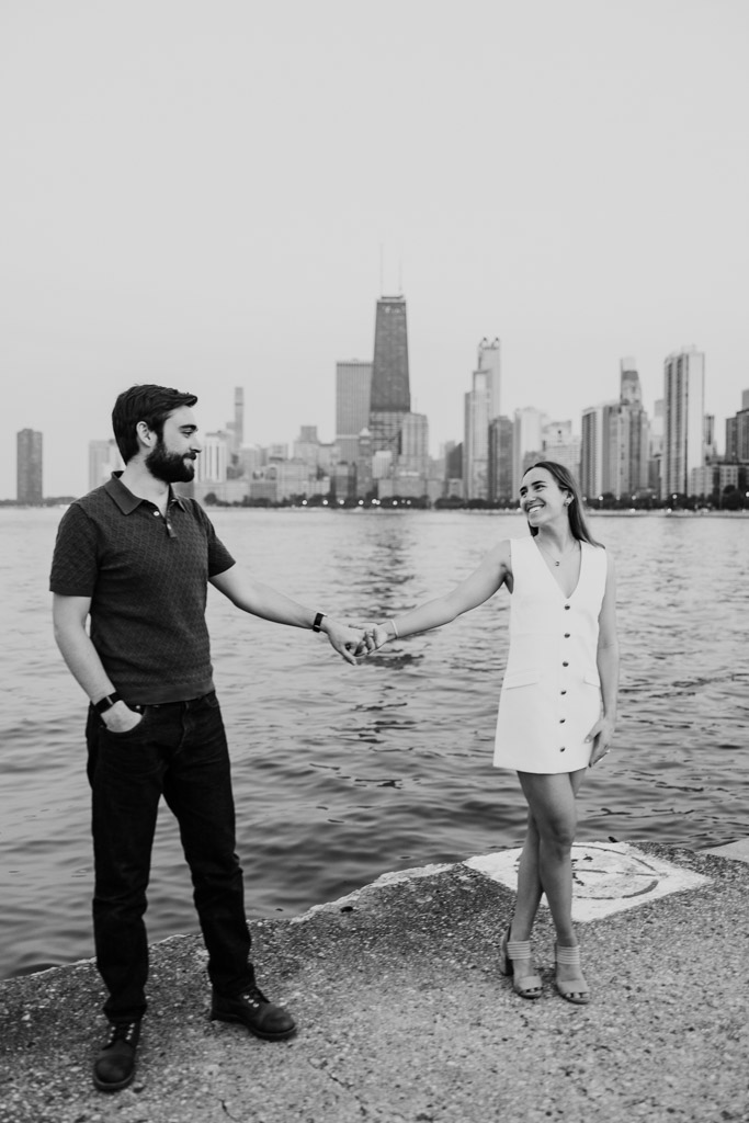 Black and white photo of couple holding hands by Lake Michigan at North Avenue Beach with the Chicago skyline in the background
