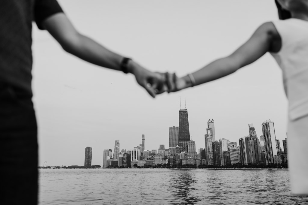 Two people holding hands with a city skyline and waterfront in the background, captured in black and white during a Chicago summer engagement.