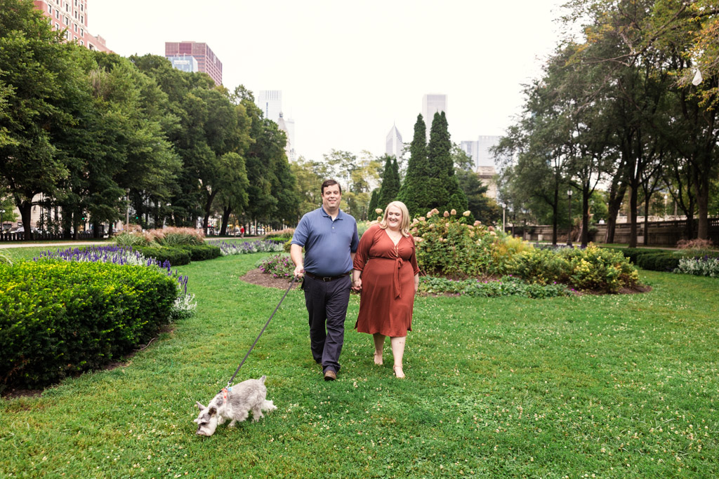 A couple walks their dog on a leash through Grant Park surrounded by trees and flowers during their South Loop engagement session