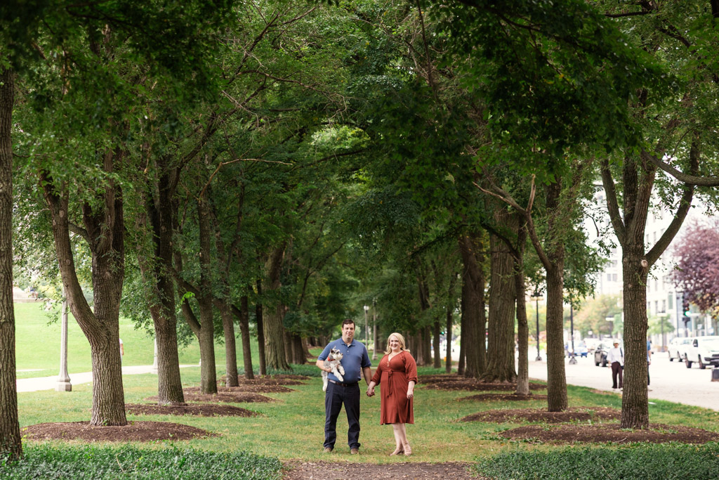 A couple holding their dog under tall trees along a path in Grant Park during their SOuth Loop engagement session