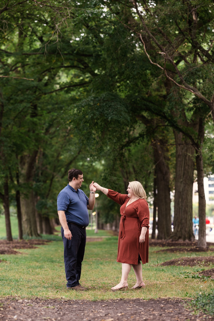 A couple dances together on a grassy path lined with tall, leafy trees in Grant Park