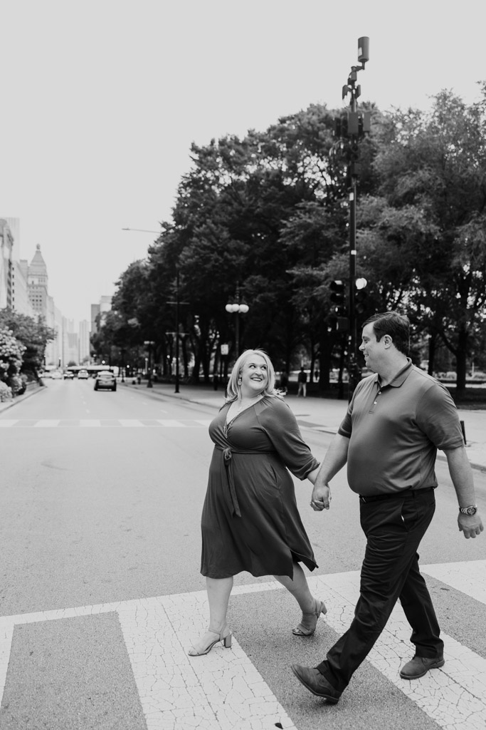 Black and white photo of couple holding hands and smiling at each other while crossing a city street during their South Loop engagement session