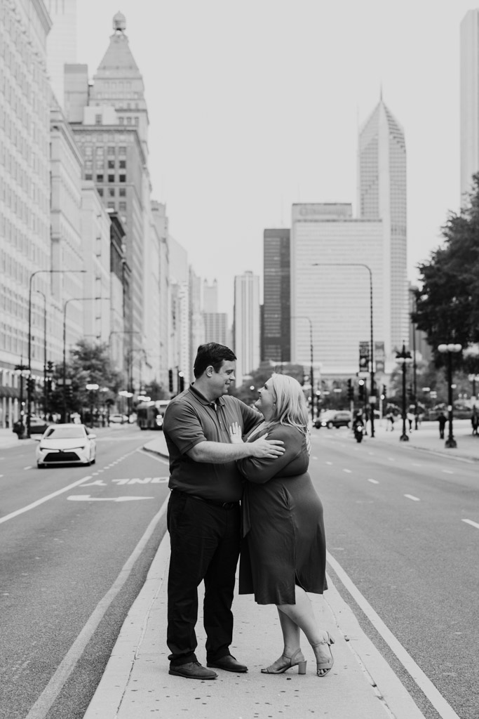 black and white photo of engaged couple embraces on a city street median in Chicago's South Loop, surrounded by tall buildings and light traffic
