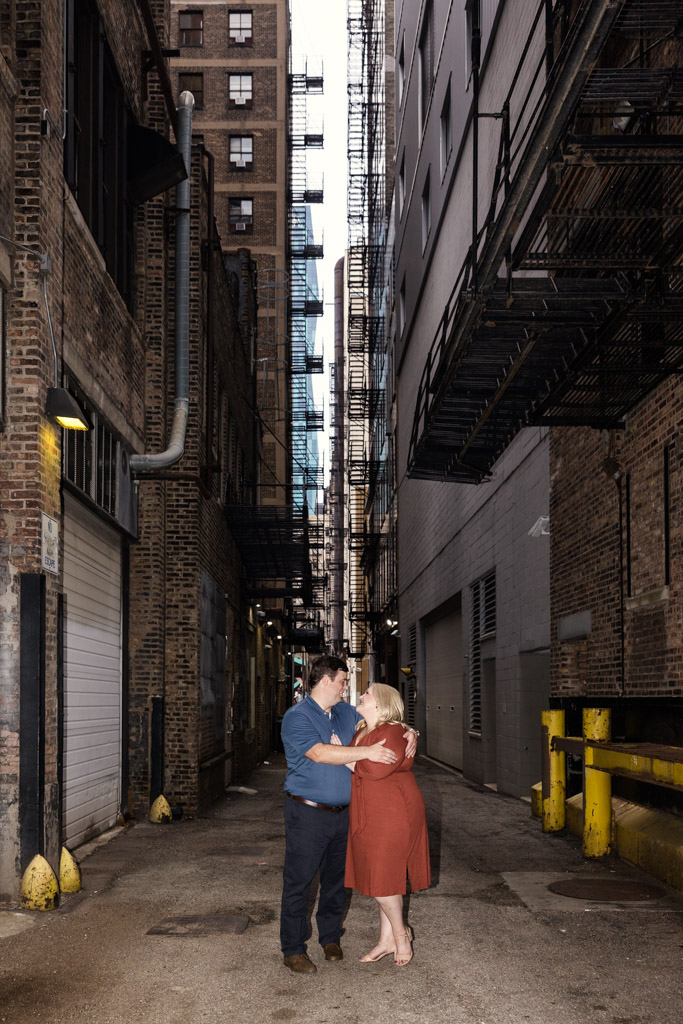 A couple holds each other in an South Loop alleyway between tall brick buildings and metal fire escapes during their downtown Chicago engagement session