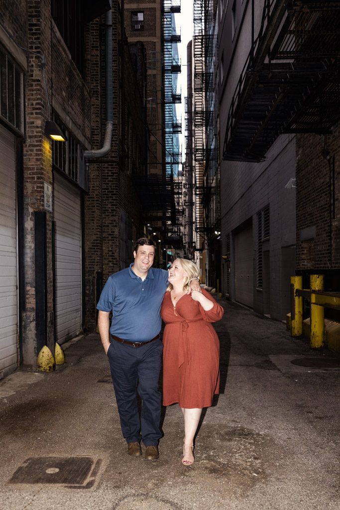 Happy couple walks arm-in-arm down a city alley in Chicago's South Loop, surrounded by brick buildings and fire escapes during their engagement session