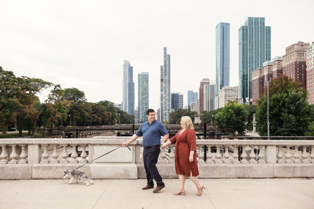 A couple enjoys a South Loop engagement stroll, walking their small dog on a leash along a city bridge with skyscrapers in the background.