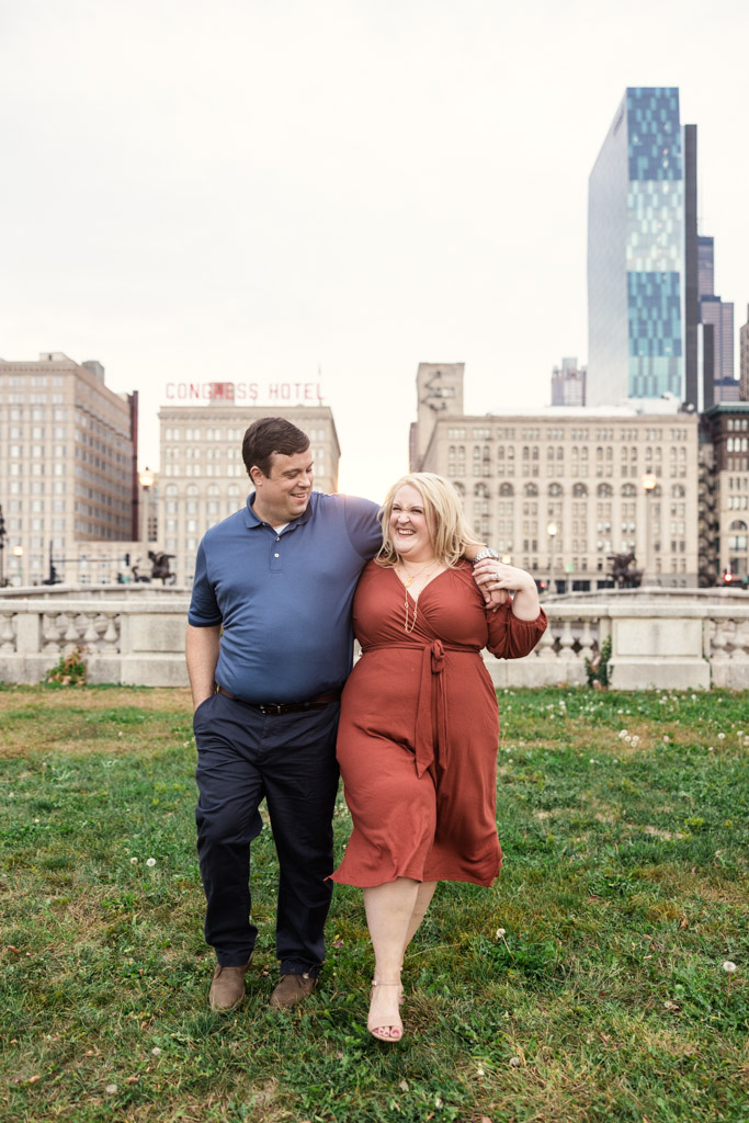 A couple smiles while walking across a grassy field in Grant Park, downtown Chicago buildings in the background