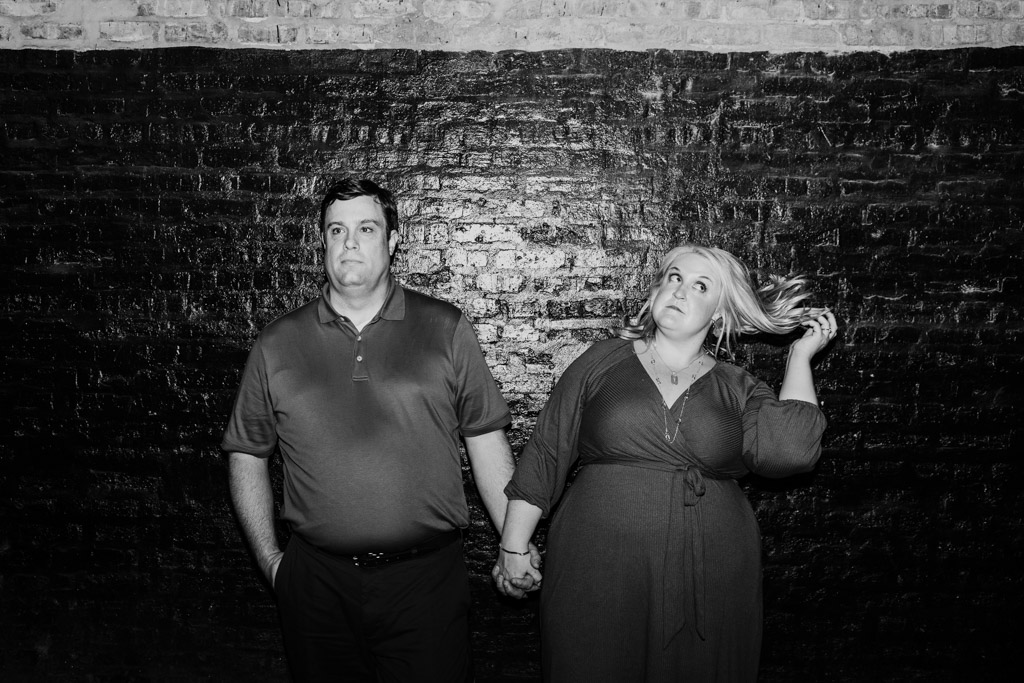 Black and white photo of engaged couple holding hands against a dark brick wall during their South Loop engagement session; the woman twirls her hair