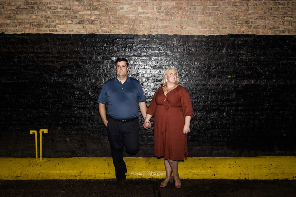 A man and woman hold hands in front of a brick wall, both looking in different directions during their South Loop engagement session