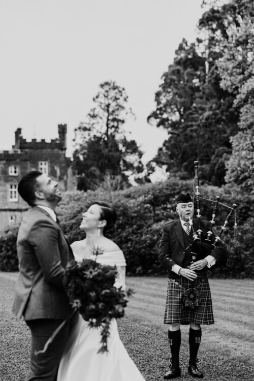 A couple laughs outdoors as a bagpiper in a kilt plays, with a castle and trees in the background.