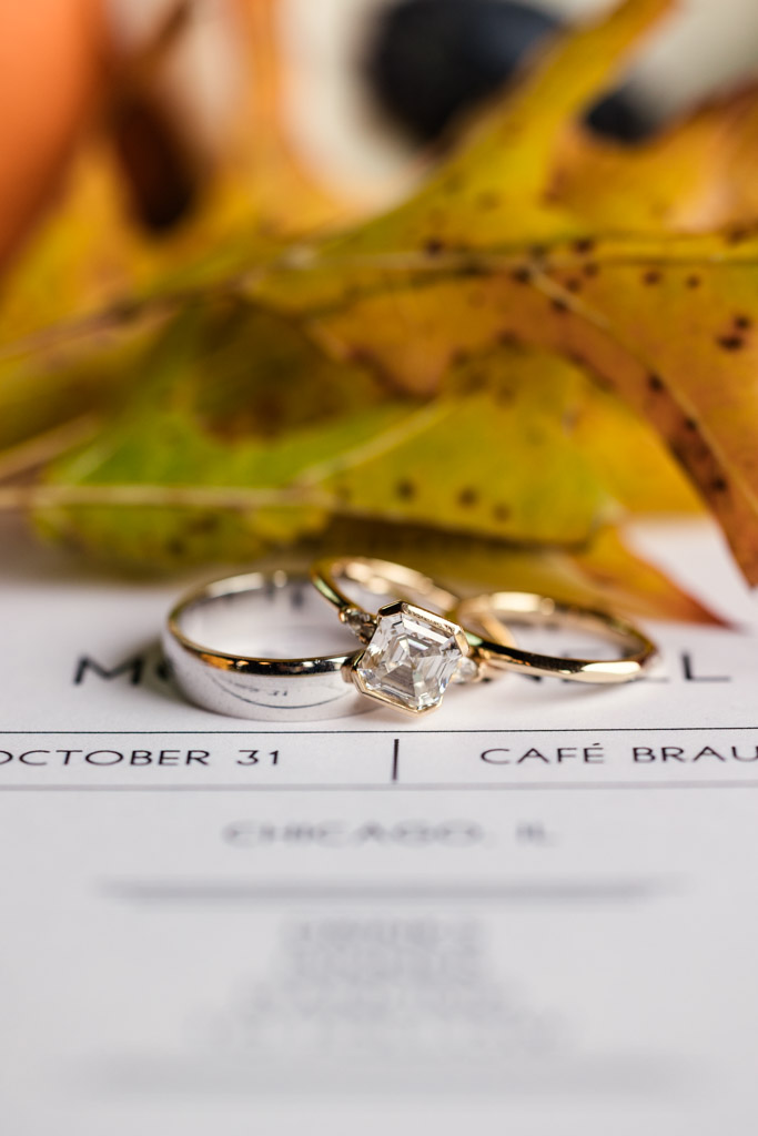 Three gold rings rest on a wedding invitation, surrounded by autumn leaves for Cafe Brauer wedding celebration