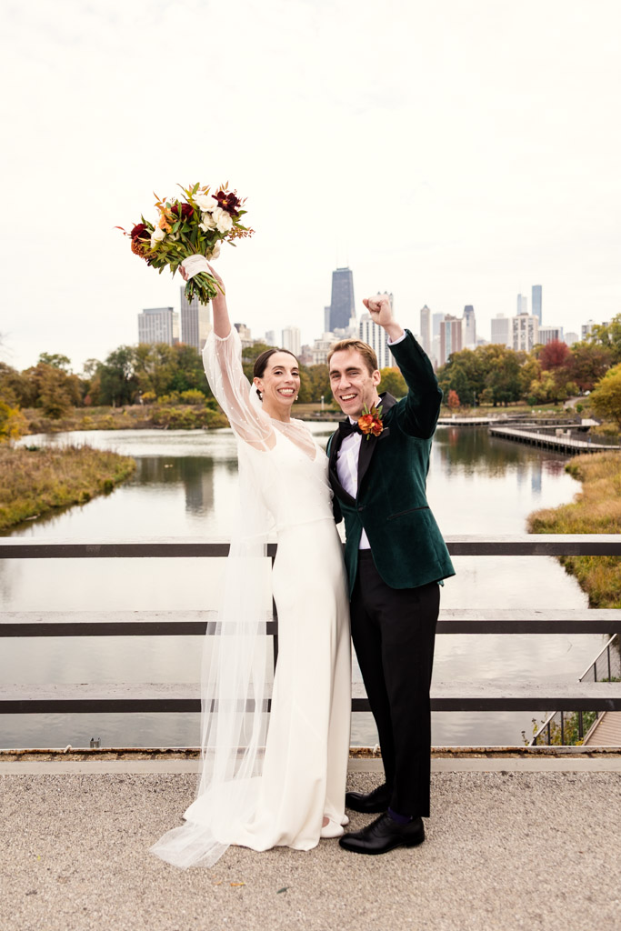 A joyful bride and groom celebrate on a Lincoln Park bridge with the Chicago skyline in the background before their Cafe Brauer wedding reception