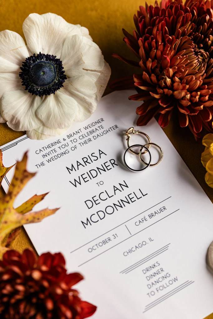 Wedding invitation with gold rings, surrounded by red, white, and yellow flowers on a yellow surface for Cafe Brauer wedding celebration