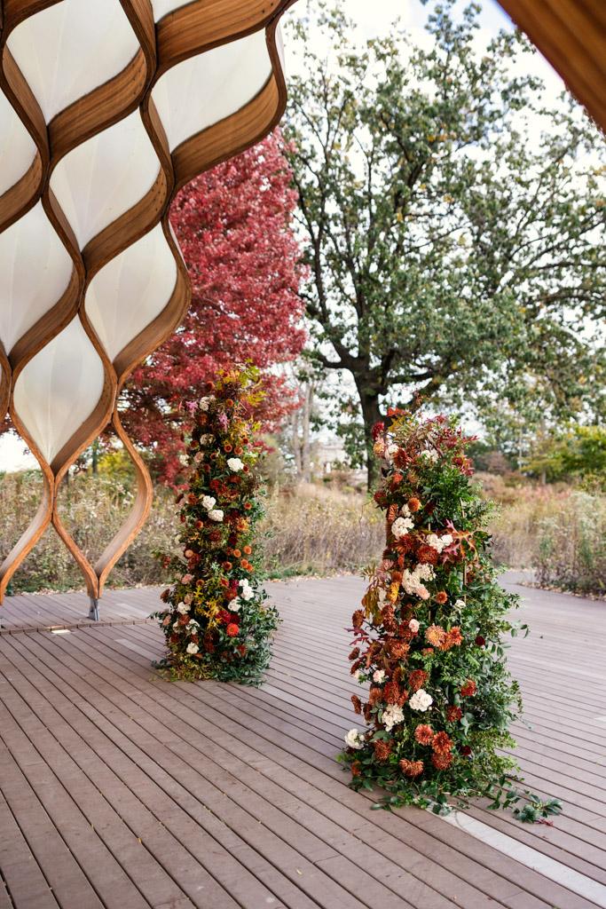 Two tall floral arrangements on a wooden deck under the Lincoln Park Honeycomb for a Cafe Brauer wedding ceremony, with autumn trees in the background