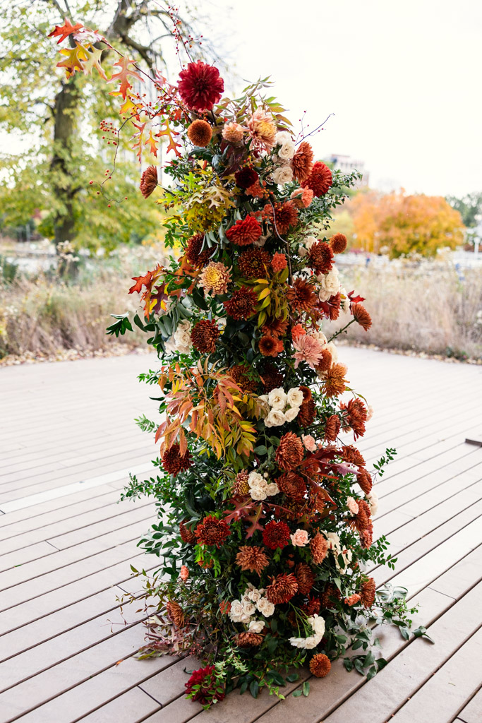 Tall floral arrangement with red, orange, and white flowers and greenery on a wooden deck outdoors for wedding ceremony at the Lincoln Park Honeycomb