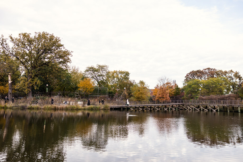 Bride walsk across a wooden bridge over a calm pond, surrounded by autumn trees and a cloudy sky