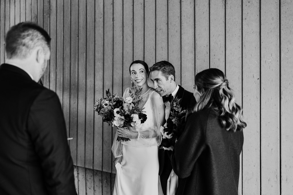 A bride and groom smile and hold bouquets while talking with two guests indoors at their elegant Cafe Brauer wedding.