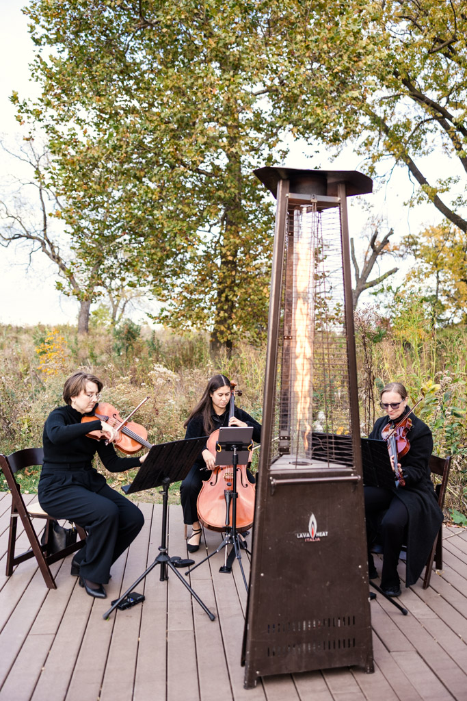 Three musicians play string instruments outdoors near a tall patio heater, surrounded by trees and foliage, during autumn Lincoln park Honeycomb wedding ceremony