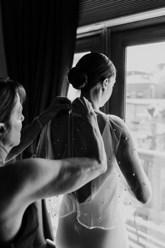 Black and white photo of woman helping bride adjust her sheer, pearl-dotted veil by a window while geetting ready for her Cafe Brauer wedding